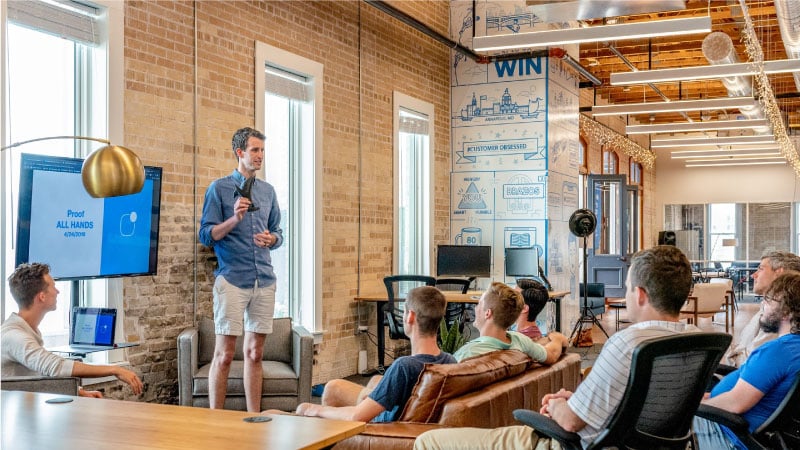 A team of employees have a meeting together in their Austin, Texas, office. One staffer is standing up in front by a large screen, giving a presentation to the rest of the team.