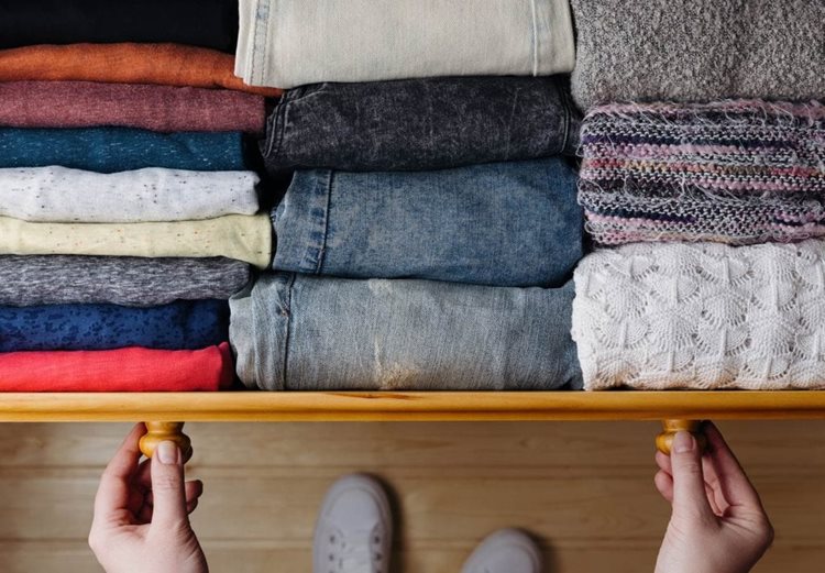 A woman's hands are opening a drawer with neatly folded shirts, jeans, and sweaters in various colors and materials.