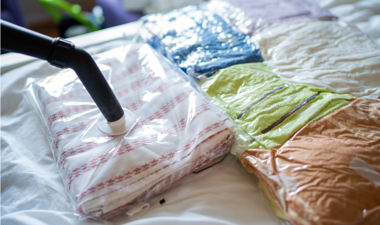 A vacuum hose is being used to suck the air out of clear vacuum-sealed bags filled with seasonal clothing of various colors.