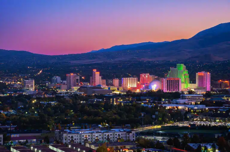 A distant view of the illuminated buildings of Downtown Reno, Nevada, at dusk. Beyond the city, the sun is setting over the mountains. The sky is colored with hues of peach and pink that fade into blues.