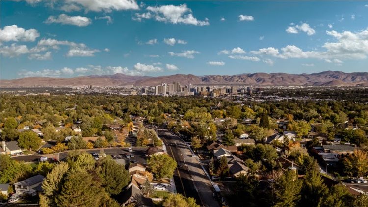 Aerial view of a residential area in Reno, Nevada, on a lovely, summer day. Single-family homes are spread out among mature trees and the city center can be seen in the distance. Beyond the city is a range of mountains.