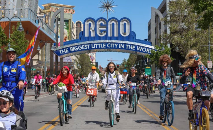 Dozens of people are riding their bikes under the Reno Arch as they participate in the Reno River Roll during Reno River Festival. It’s a bright day and some of the participants are wearing fun costumes.