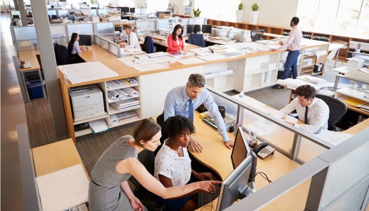A man and a woman are helping another woman with something on her computer. In the adjoining cubicle, another man is writing something down. There are a few other coworkers in the background, working on different projects.