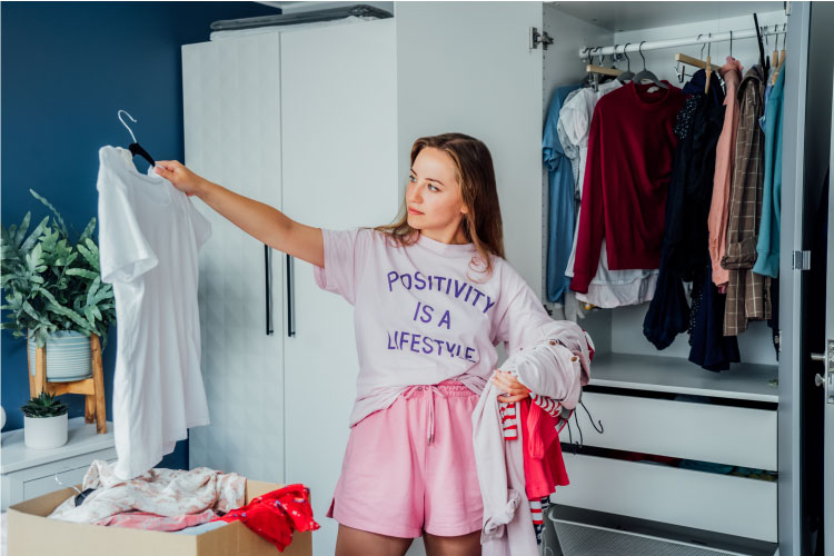 A young woman is wearing a shirt that says, “Positivity is a lifestyle.” She’s going through the clothes in her closet, choosing what to keep and what to get rid of.