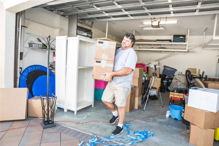 A home decluttering professional is carrying two boxes out of a cluttered garage.