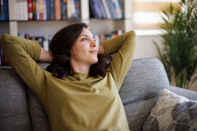 A young woman is relaxing on her couch after decluttering her home. The space feels more peaceful and calm, and it’s easier for her to relax now that there’s no more clutter.