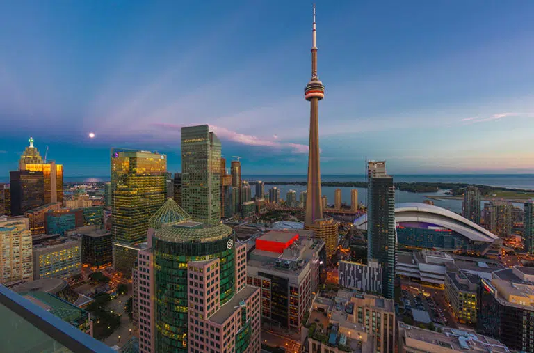 Aerial view of Toronto city centre at dusk. There are a variety of large structures and buildings of different styles and heights. The moon can be seen hanging low in the sky, and the last glimpses of sunset are fading on the horizon.
