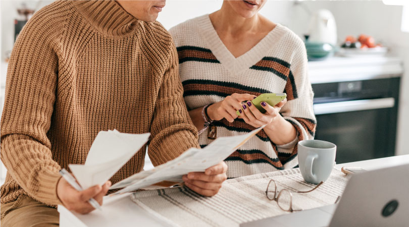 A husband and wife sit together at their laptop, reviewing their bills and cost of living expenses.