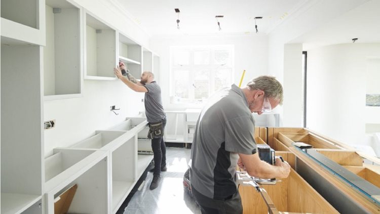 Two contractors in gray shirts work to install new kitchen cabinets in a residential home. One is working on the kitchen island while the other is installing an upper cabinet in the back.