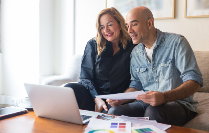 A happy couple looks over their remodeling plans on a laptop. They also have sheets of paper with paint color samples printed on them. 