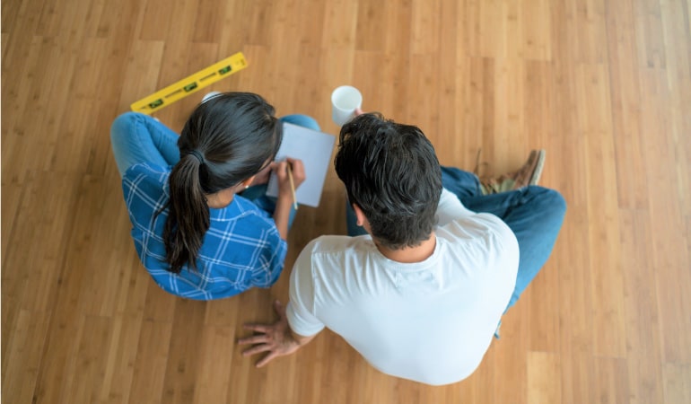 A couple sitting together on the floor of their home beside a level tool. They’re making a home renovation checklist to track the steps for their upcoming home renovation.