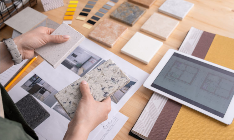 A woman looks over paint and countertop samples, holding two countertop samples in her hands, as she considers how she would like to remodel her kitchen.