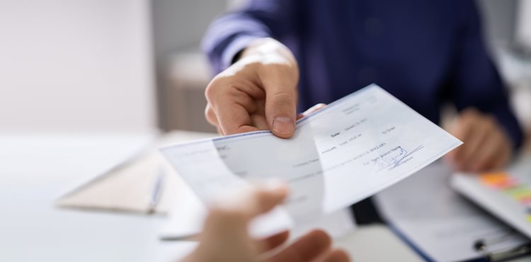 A first-person view of someone reaching out to receive a paycheck from an employer. The man handing the check across the desk is wearing a blue button-up shirt, but his face is not visible. 