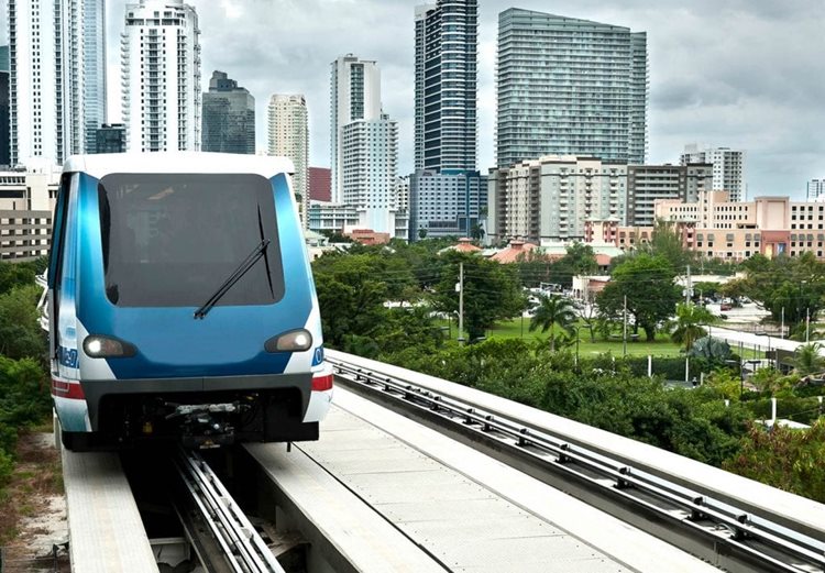 A head-on view of the Miami Metromover tram as it travels on the tracks with the Miami city skyline visible behind it. Below the tracks, palm trees and other lush foliage are visible, as well as pastel-colored buildings, typical of Miami.