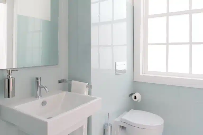 A half bathroom, featuring a simple blue and white color palette and a modern white ceramic toilet and sink. Natural light is streaming in from the large window above the toilet.