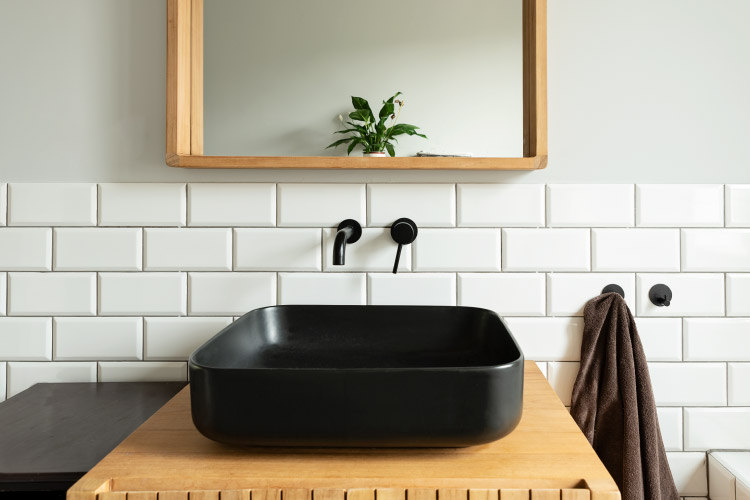 A half bath counter, sink, and mirror. The sink is a modern black stone basin in a rounded square shape. The faucet is installed directly into the white subway tiles on the wall behind the sink. 