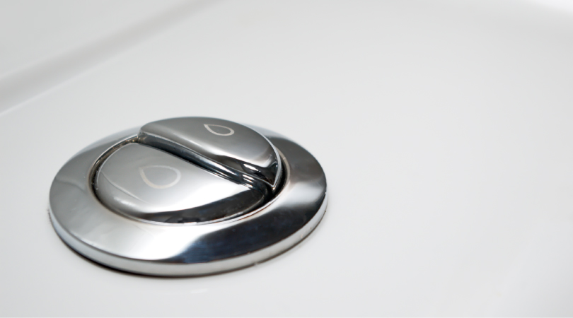 A close-up view of metallic, dual-flush toilet buttons. The right button has a small water drop etched on it, and the left button has a larger water drop etched on it, indicating increased water flow.