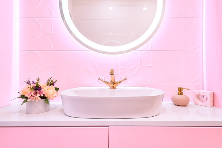 A white bathroom sink and counter are accented by a matching pink vanity and tile wall. Above the sink hangs a circular light-up vanity mirror, while flowers, a soap dispenser, and a small heart statue decorate the countertop. 