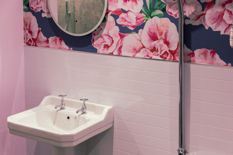 A white pedestal sink with two faucets sits beneath a simple white mirror in a half bath. The bottom half of the wall features white subway tiles, and the top half is covered in bold floral wallpaper. 