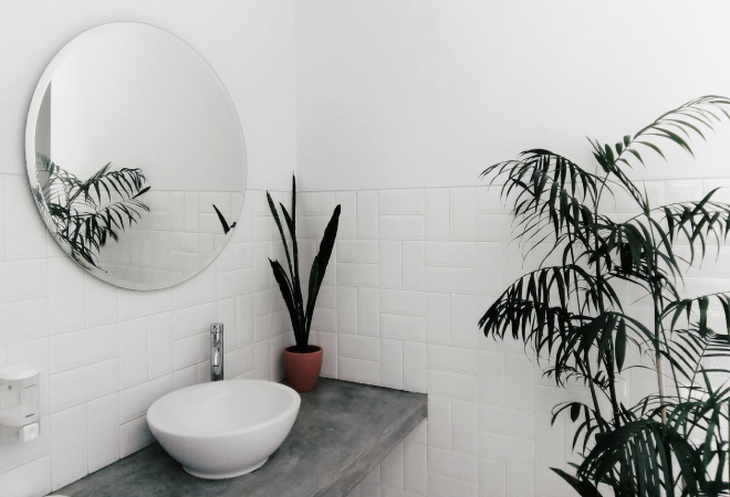 A white tiled half bathroom with a concrete counter, a ceramic bowl sink, and a round, frameless mirror. There are plants throughout the bathroom, as well.