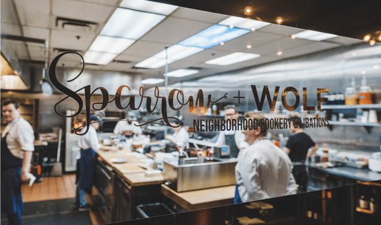 View inside the Sparrow + Wolf kitchen in Las Vegas, Nevada, through a glass window. The window is decorated with a metallic gold decal that reads “Sparrow + Wolf Neighborhood Cookery & Libations.” The chef and other cook staff are manning their stations inside the organized kitchen.
