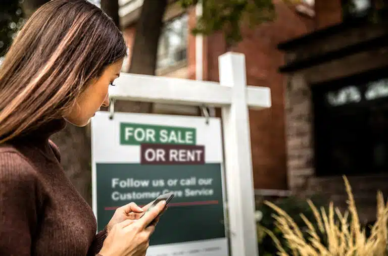 A woman is using her mobile phone to contact the realtor listed on a “for sale or rent” sign in front of a home