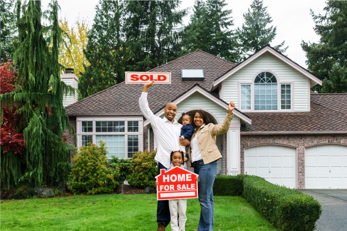 A happy family of four is standing in front of their new home. The older daughter is holding a “home for sale” sign and the father is holding a “sold” sign.