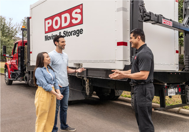 A young couple stand in front of a PODS container as their driver explains how their things will be shipped to their next home