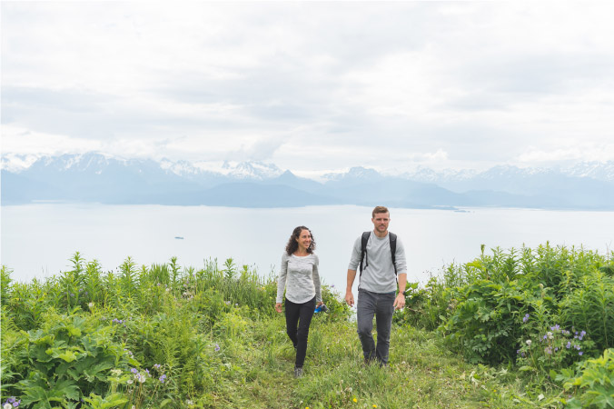 A young couple is hiking in Alaska with a view of the water and mountains in the distance