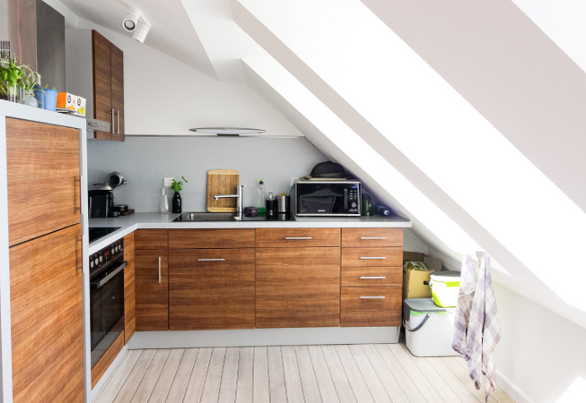 A small attic that’s been renovated into an extra kitchen space with a stove top, oven, sink, and wooden cabinets. The white walls contrast beautifully with the natural wood cabinetry.