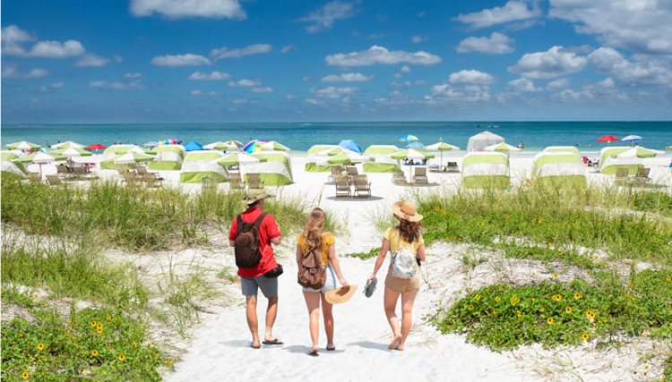 Three adults are walking through sand dunes at Clearwater Beach in Florida, toward the blue waters of the Gulf of Mexico.