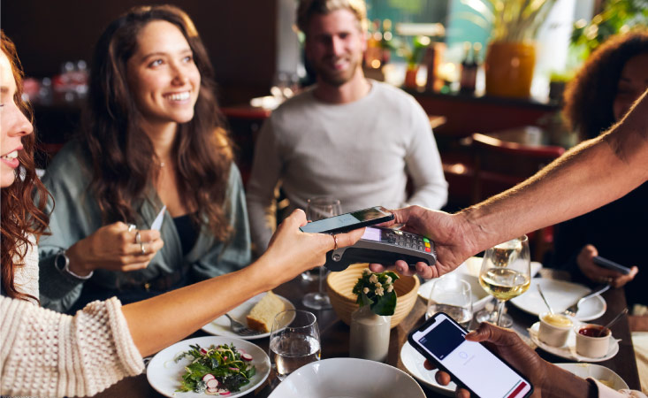 Five friends are sitting at a table in a restaurant, taking turns paying for their part of the meal using mobile phones or credit cards.