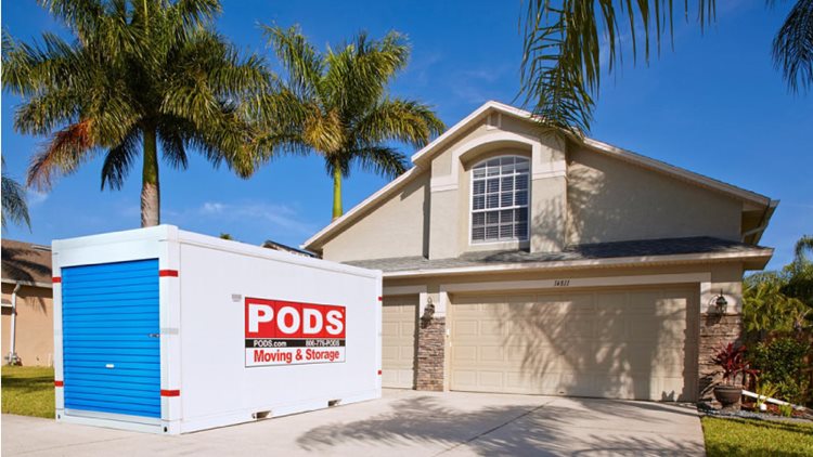 A PODS portable moving and storage container is positioned in the driveway of a residential home in St. Petersburg, Florida. The home has a stucco and brick exterior and there are a couple of tall palm trees beside it.