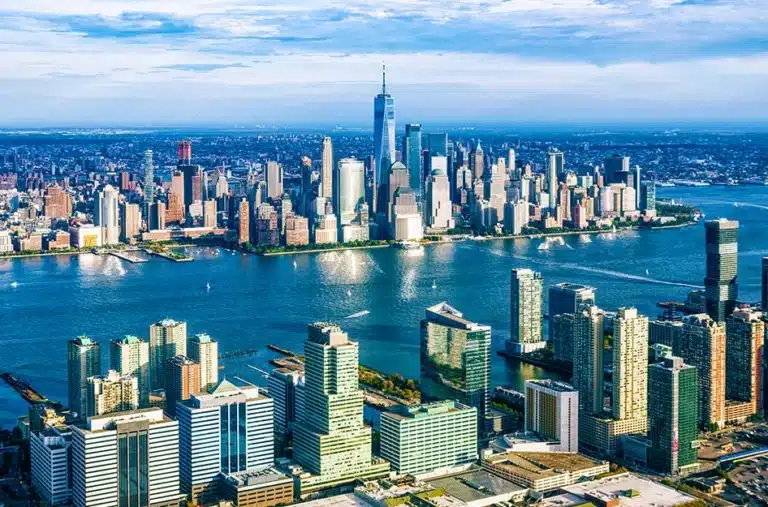 The New Jersey City coastline, looking out toward New York City. Skyscrapers on both sides of the water tower over smaller nearby buildings.