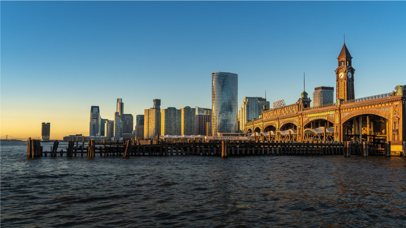 The Hoboken, New Jersey, waterfront at dusk. A pier in partial disrepair is jutting out into the water as the last of the day’s sunlight reflects off city buildings. 