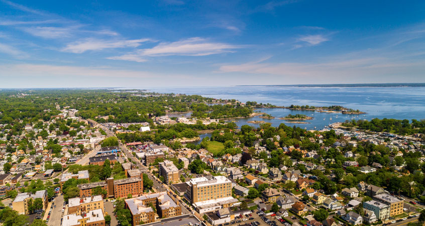 Aerial view of the city of New Rochelle, New York, and its coast on a sunny day. Square-shaped brick buildings make up the city center, and residential homes and mature trees fill the surrounding areas.