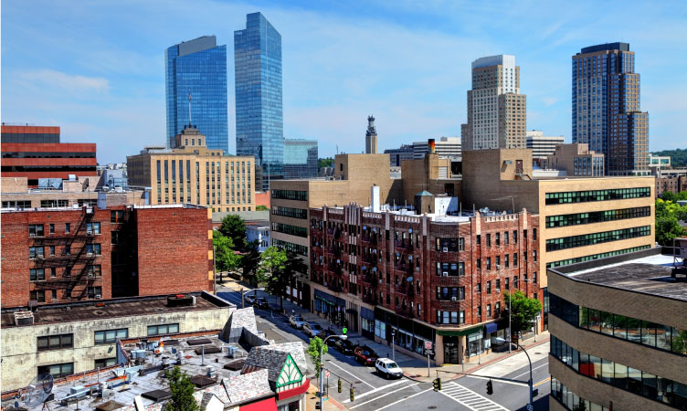 Aerial view of downtown buildings in White Plains, New York, on a sunny day. Cars are parked along the city street below, and the blue sky is filled with faint wisps of clouds.