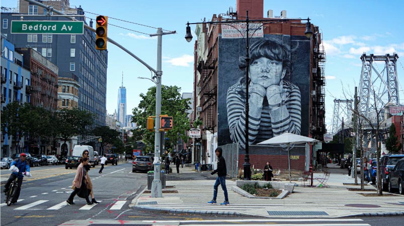 Pedestrians crossing Bedford Avenue in Williamsburg, New York, on a clear day. A large black-and-white mural titled “Lost Time,” depicting a young girl with her chin resting in her hands, decorates a brick building in the middle of the scene. The Williamsburg Bridge can be seen in the background. 