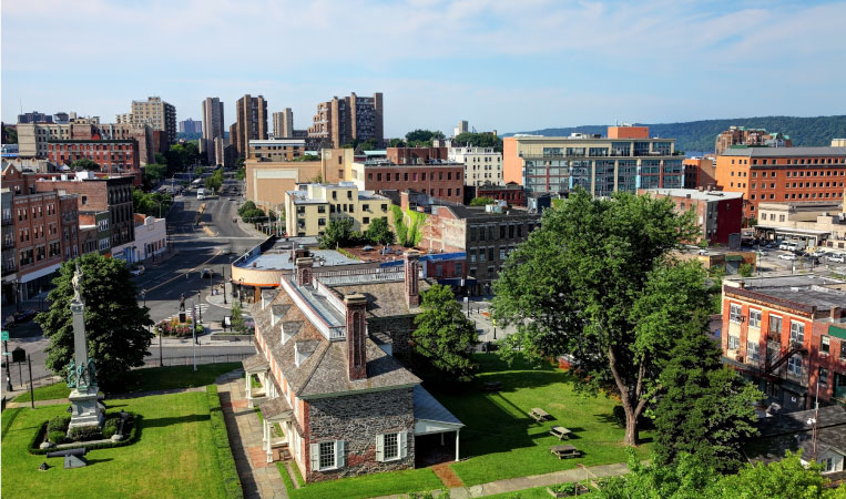 Aerial view of the city of Yonkers, New York, on a bright summer day. The foreground features greenspaces and mature trees, and a low mountain range can be seen in the distance. 
