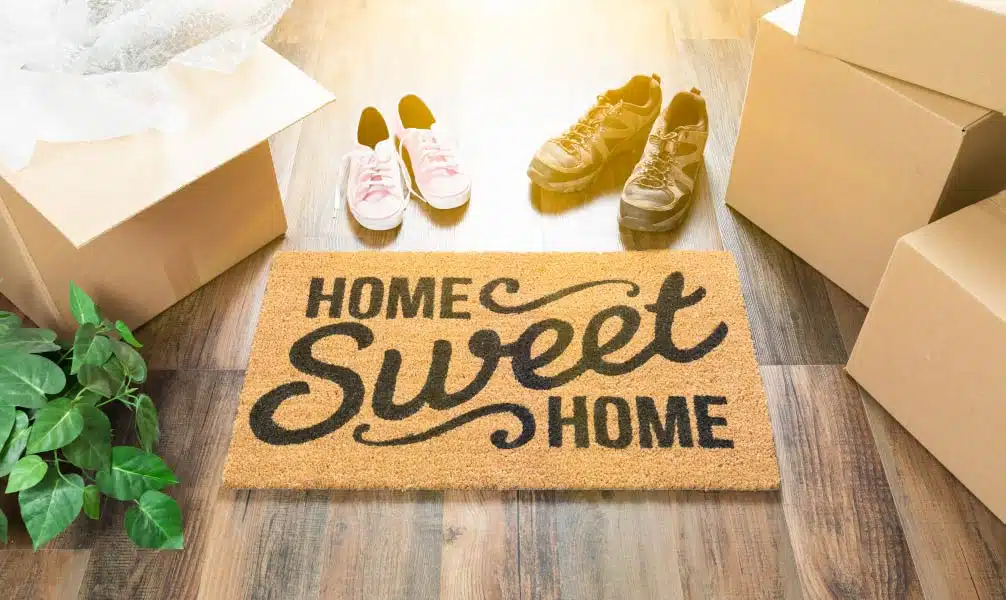 Morning light is shining on several moving boxes, two pairs of sneakers, and a “Home Sweet Home” doormat in the entryway of a new home.