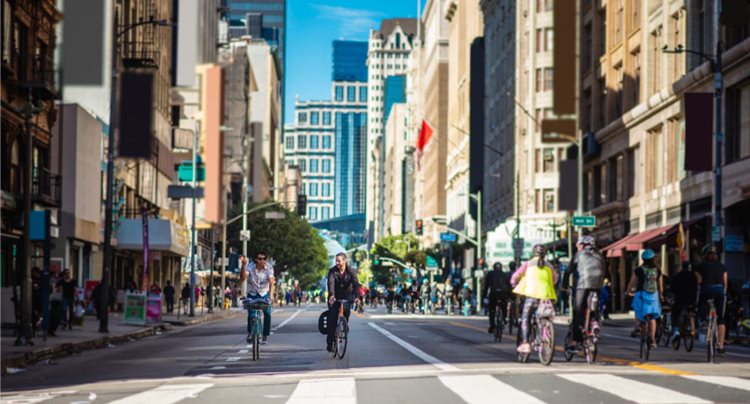 Angelenos cycle through sunny Downtown Los Angeles between tall city buildings.