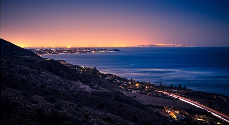 View of the Pacific Coast Highway and the hills in Malibu seen at night. The road is lit up with streams of headlights, and the glow of a city can be seen in the distance.
