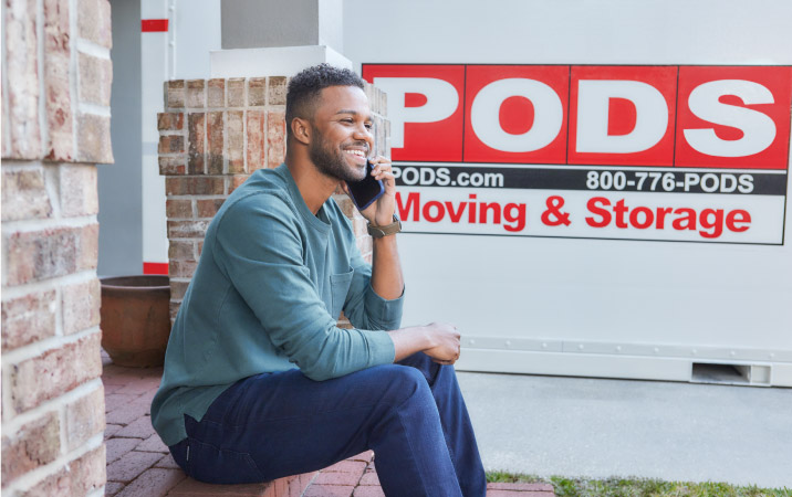 A man is smiling while he sits on his porch and talks on his cell phone. Behind him, in the driveway, is a PODS portable moving and storage container, which he is using to move to one of the best Los Angeles neighborhoods.