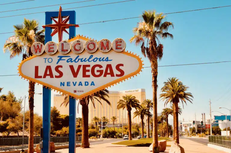 The colorful “Welcome to Fabulous Las Vegas Nevada” sign is positioned between a tall pair of palm trees beside a road in Las Vegas, with the Mandalay Bay hotel visible in the background.