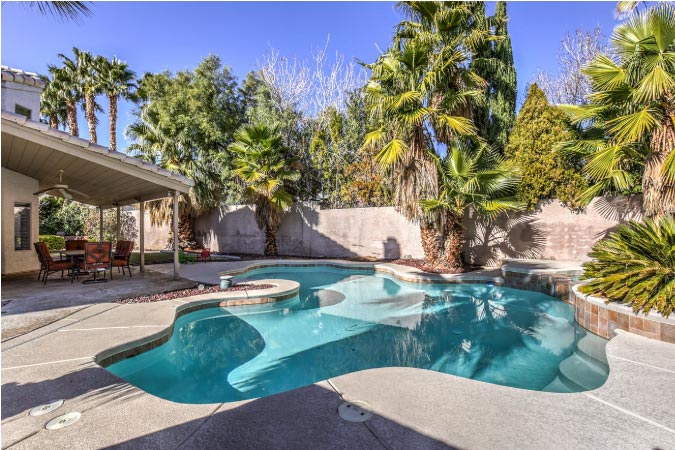 The backyard of a home in Peccole Ranch, Las Vegas, featuring a custom in-ground pool, covered patio, and various palms.