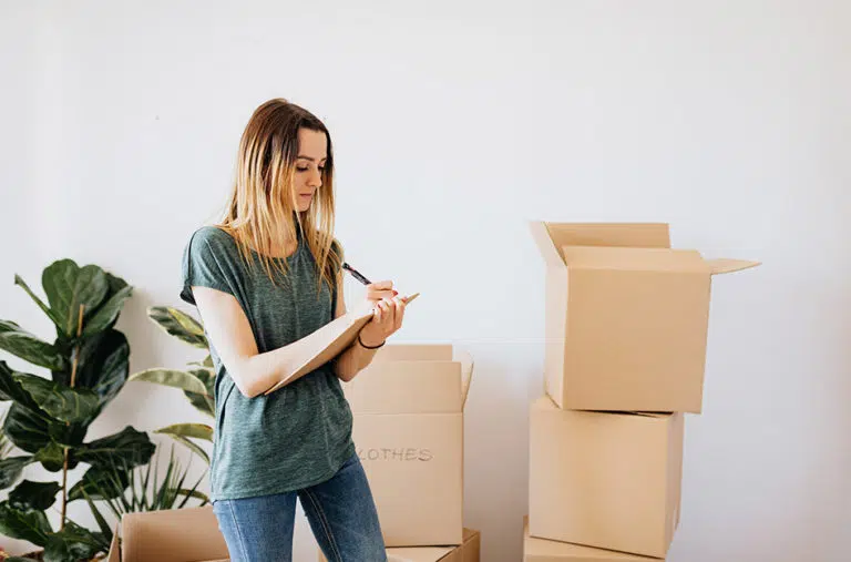 A woman standing beside stacked moving boxes, making an inventory of the things she’s packing inside them. There are a few large houseplants beside her.