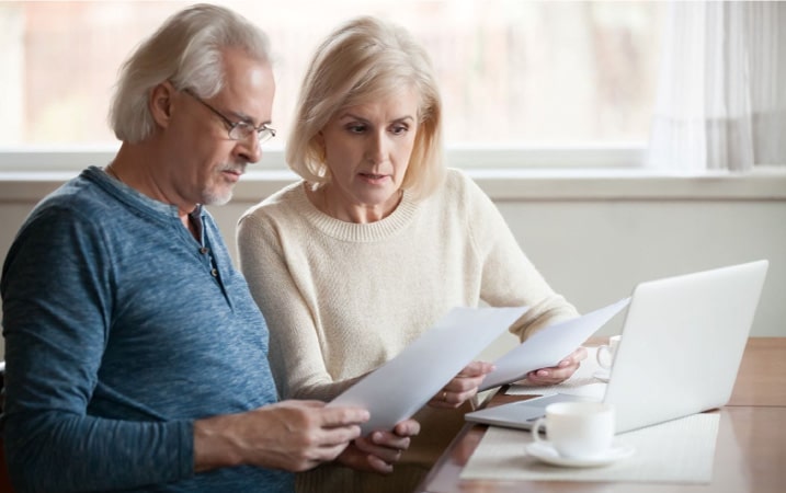 An older couple sit down at a compter to review renters and homeowners insurance. They are seated at a white laptop with two cups of coffee in white cups.