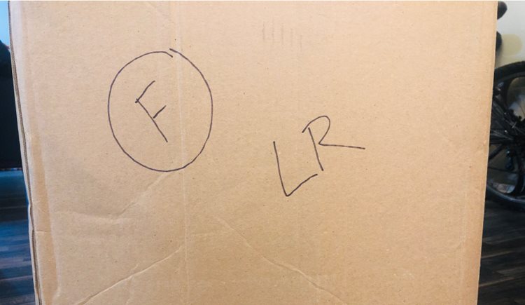 The side of a moving box with handwritten shorthand labels indicating that it is fragile (a big “F” with a circle around it) and packed with items from the living room (a big “LR”).