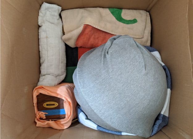 View from above, looking into an open cardboard box. In the box, various items are wrapped in clothing and other linens and packed snuggly together. This is a handy way of avoiding the need to use bubble cushioning roll while also packing more of your things in a single moving box.