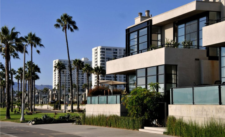 A modern Los Angeles apartment building with concrete and glass elements. There’s decorative landscaping in the front of the building, a number of tall palm trees, and other larger apartment buildings in the background.